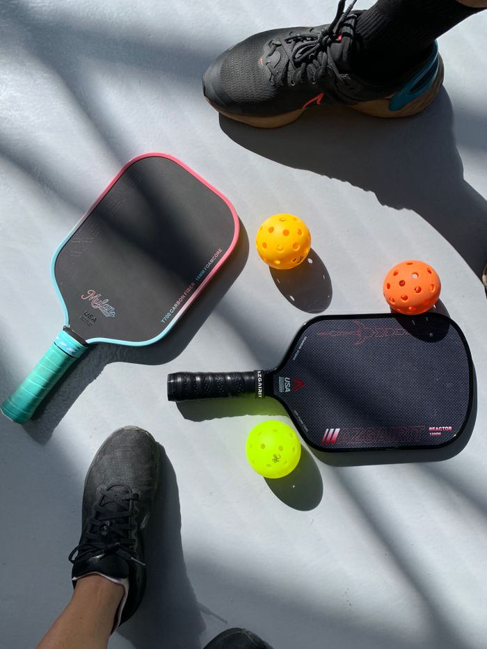 services-02 Two pickleball paddles and balls on a sunlit court, with players' feet showing.